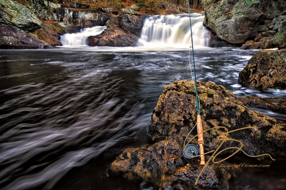 A Sage 1 weight fly rod with an Abel TR Light reel, leening up against a stream side rock, with a waterfall as background. Long exposure giving the water going over the falls a smooth, silky look.