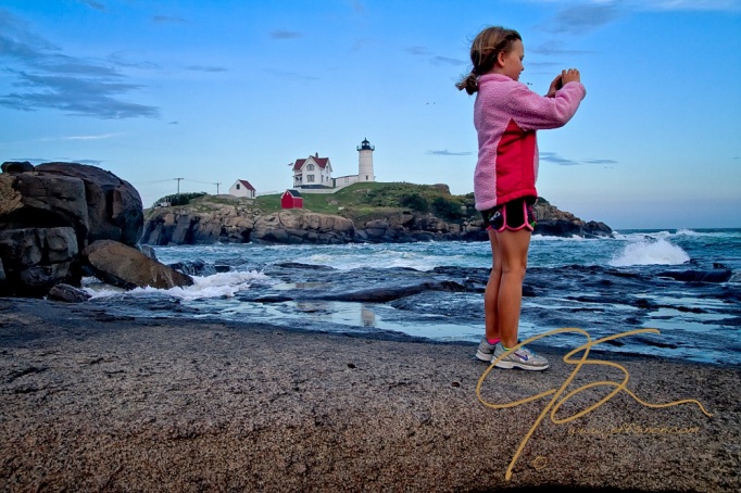 With the Cape Neddick, aka "Nubble" lighthouse in the background over the rough windswept sea, a young girl dressed in shorts and a pink fleece jacket, stands on the rocks and takes a photograph with her iPhone. Blonde hair pulled back in a ponytail without a care in the world.