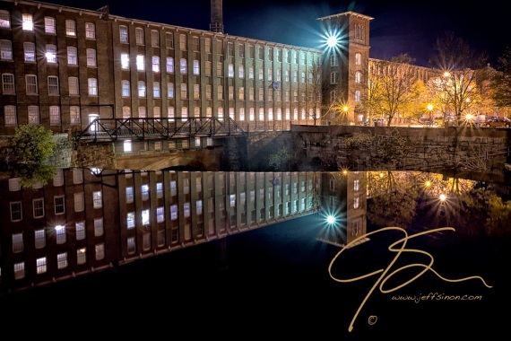 The Cocheco Mill Building in downtown Dover, NH. The bright blue-white spotlight on the tower shining brightly, a mirror image of the building reflected in the glass smooth water above the waterfalls. Numerous widows are lit from within in this long brick structure.